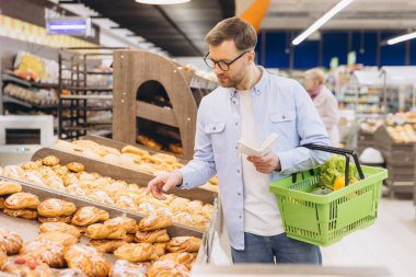 Man choosing pastry from a shelf, holding a shopping basket and a grocery list in supermarket bakery