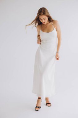 Young woman wearing elegant white dress and black high heels posing on white background in studio, looking down