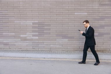 Young manager using mobile phone while walking on the street next to a brick wall