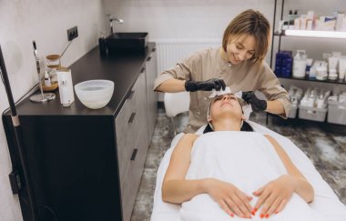 Beautician wearing black gloves cleaning face of customer lying on massage table in beauty salon