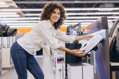 Young curly haired woman choosing a new robotic vacuum cleaner in an appliance store, smiling and looking at the product