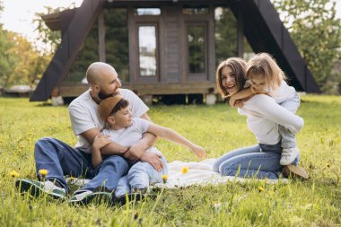 Smiling family having fun sitting on blanket in the grass in front of their modern a frame house