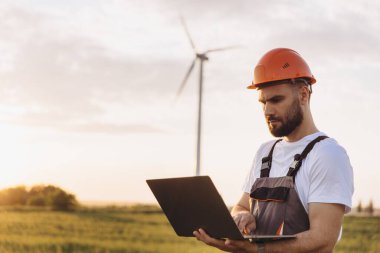 Engineer wearing overalls and hardhat is using a laptop in front of a wind turbine at sunset, working on renewable energy production