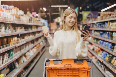 Smiling woman using her phone and credit card while making purchases for groceries in a bright, organized supermarket aisle