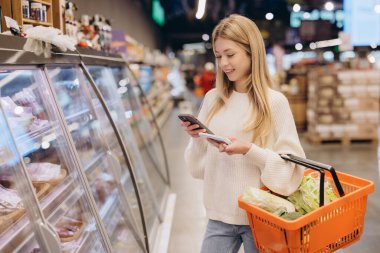 Akıllı telefonunu ve alışveriş listesini kullanan genç bir kadın markette soğutulmuş rafların yanında yiyecek seçiyor.
