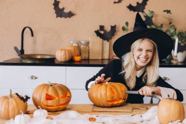 Young woman with a witch hat happily carving pumpkins for a Halloween party in a cozy kitchen, embracing festive autumn traditions