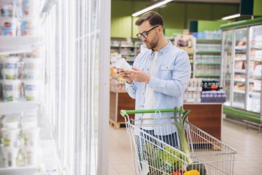 Man pushing shopping cart and reading food label in supermarket refrigerator