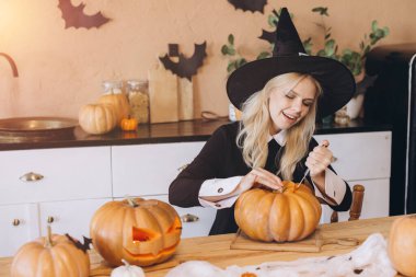Young woman wearing witch hat carving pumpkin for Halloween party in kitchen decorated with bats and pumpkins