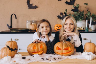 Mother and daughter smiling and joyfully carving pumpkins together in a kitchen adorned with festive Halloween decorations