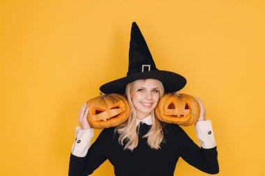 Young blonde woman wearing witch costume holding two carved pumpkins on yellow background for Halloween