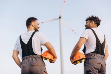 Two engineers holding helmets and admiring a wind turbine in the background, celebrating sustainable energy