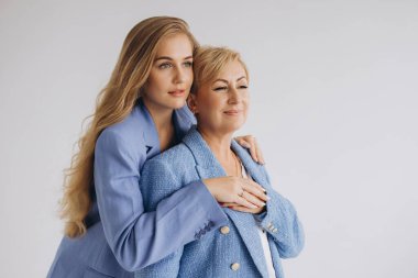 Smiling businesswomen mother and daughter wearing blue suits are hugging and looking away on white background
