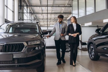 Car salesman presenting a new vehicle to a female customer inside a modern car dealership, engaging in discussion about purchasing options