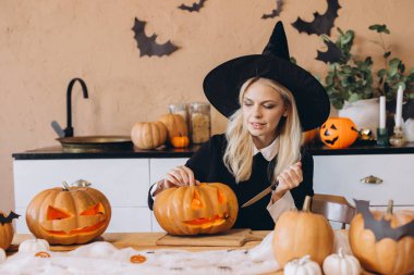 Young woman dressed in a witch costume, skillfully carving a pumpkin for Halloween with a knife in her cozy kitchen, creating festive decor