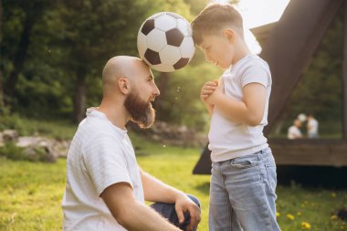 Father and son joyfully balancing a soccer ball between their heads, celebrating Father's Day together in a sunny park filled with laughter