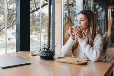 Freelancer enjoying a relaxing moment with a cup of tea in a modern cafe, laptop and smartphone resting on the table beside her