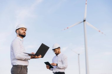 Two engineers are inspecting wind turbines with a laptop and a tablet, ensuring efficient and sustainable energy generation