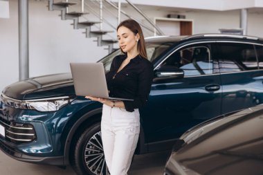 Saleswoman using laptop while standing next to new car in modern showroom