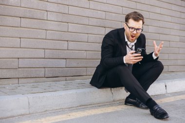 Frustrated businessman in suit and tie screaming at his phone while sitting on a street curb
