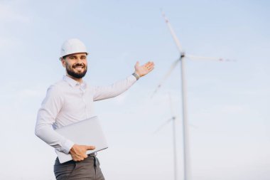 Smiling engineer with laptop and hardhat showing wind turbines in a wind farm for sustainable energy production