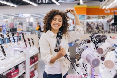 Smiling woman with curly hair testing a new hair dryer in an electronics store, enjoying the experience while touching her hair