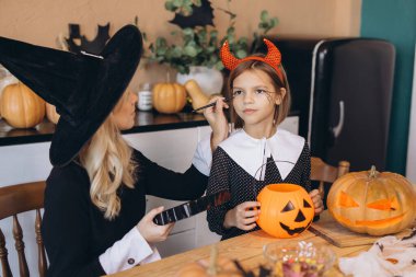 Mother wearing witch hat applying Halloween makeup on her daughter's face, sitting at wooden table with pumpkins and candies