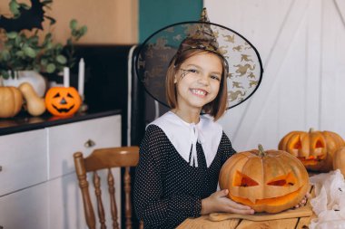 Young girl wearing witch hat and halloween costume holding carved pumpkin celebrating Halloween at home