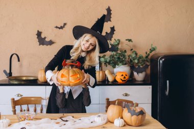 Mother wearing witch costume is helping her daughter to hold a carved Halloween pumpkin in front of her face
