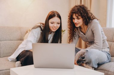 Two teen students working together on a school project using a laptop, sitting on a sofa in the living room