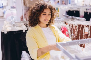 Smiling curly haired woman shopping for clothes in a fashion store, looking at a rack of pants and shirts