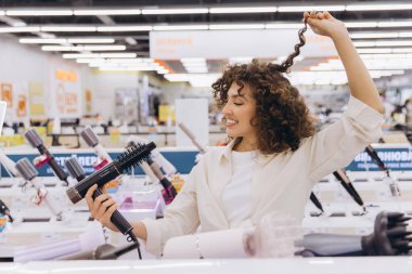 Smiling curly haired woman testing new hair dryer in appliance store, enjoying shopping and beauty products