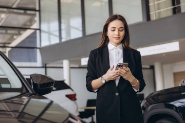 Young saleswoman using mobile phone app, choosing and comparing cars at modern dealership showroom