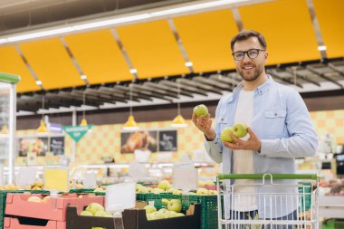 Male shopper selecting ripe apples in supermarket produce section, grasping fresh produce with cheerful expression