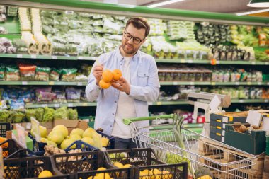 Customer buying oranges in grocery store, holding a bag of oranges, shopping cart visible in background