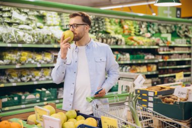 Customer smelling a green apple while holding a shopping list and pushing a cart in a supermarket