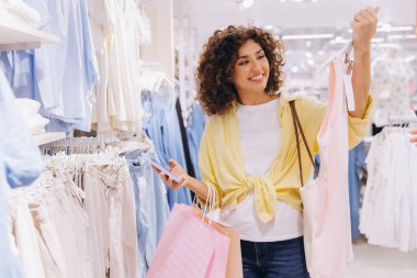 Smiling curly haired woman holding clothes and shopping bags, enjoying shopping spree in clothing store