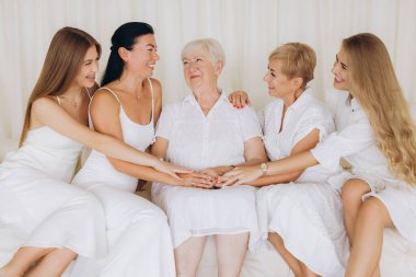 Four generations of women, wearing white dresses, sitting on a white couch, holding hands, smiling and laughing, celebrating their family bond