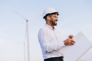 Young adult engineer holding blueprint inspecting wind turbine in a field for sustainable energy generation
