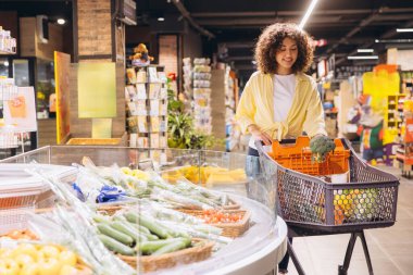 Customer selecting fresh broccoli while grocery shopping in a brightly lit supermarket, filling her basket with healthy vegetables
