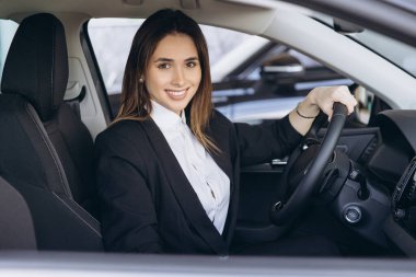 Saleswoman sitting in new car and holding steering wheel, smiling and looking at camera in car dealership showroom