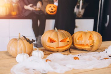 Halloween pumpkins carved with spooky faces sit on a wooden table, illuminated by warm light, with family members preparing for the festivities in the blurred background