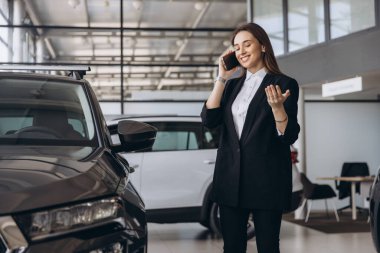 Smiling saleswoman engaging in a phone conversation while presenting a new car to an interested customer in a modern dealership showroom