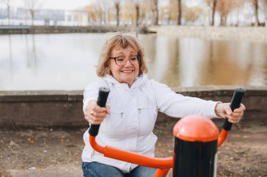 Happy senior woman exercising using outdoor gym equipment in a park near a lake, promoting healthy lifestyle and active aging