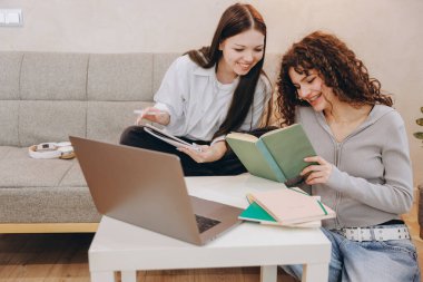 Two smiling students learning and preparing exam using laptop, tablet and book sitting on sofa at home