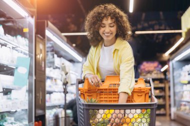Smiling curly haired woman pushing shopping cart and choosing fresh fruits and vegetables in supermarket