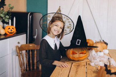 Little girl wearing witch hat smiling and posing with carved pumpkins and Halloween decorations on a wooden table in kitchen