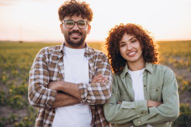 Interracial agronomists couple smiling with arms crossed in cultivated soybean field at sunset