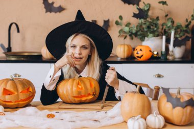 Blond woman wearing witch hat carving jack o' lantern from pumpkin with knife in decorated kitchen for Halloween party