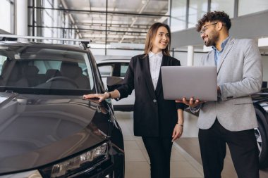 Car salesman holding a laptop is presenting a new car to a businesswoman at the dealership, discussing features and financing options