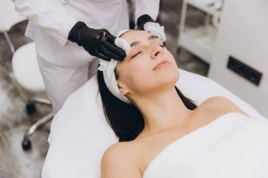 Beautician wearing black gloves is applying skincare product on a woman's face with cotton pads, in a professional beauty salon